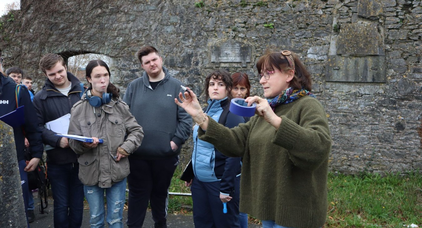 Instructor in green jumper stands before group of students showing how masking tape and marker are used to number the gravestones