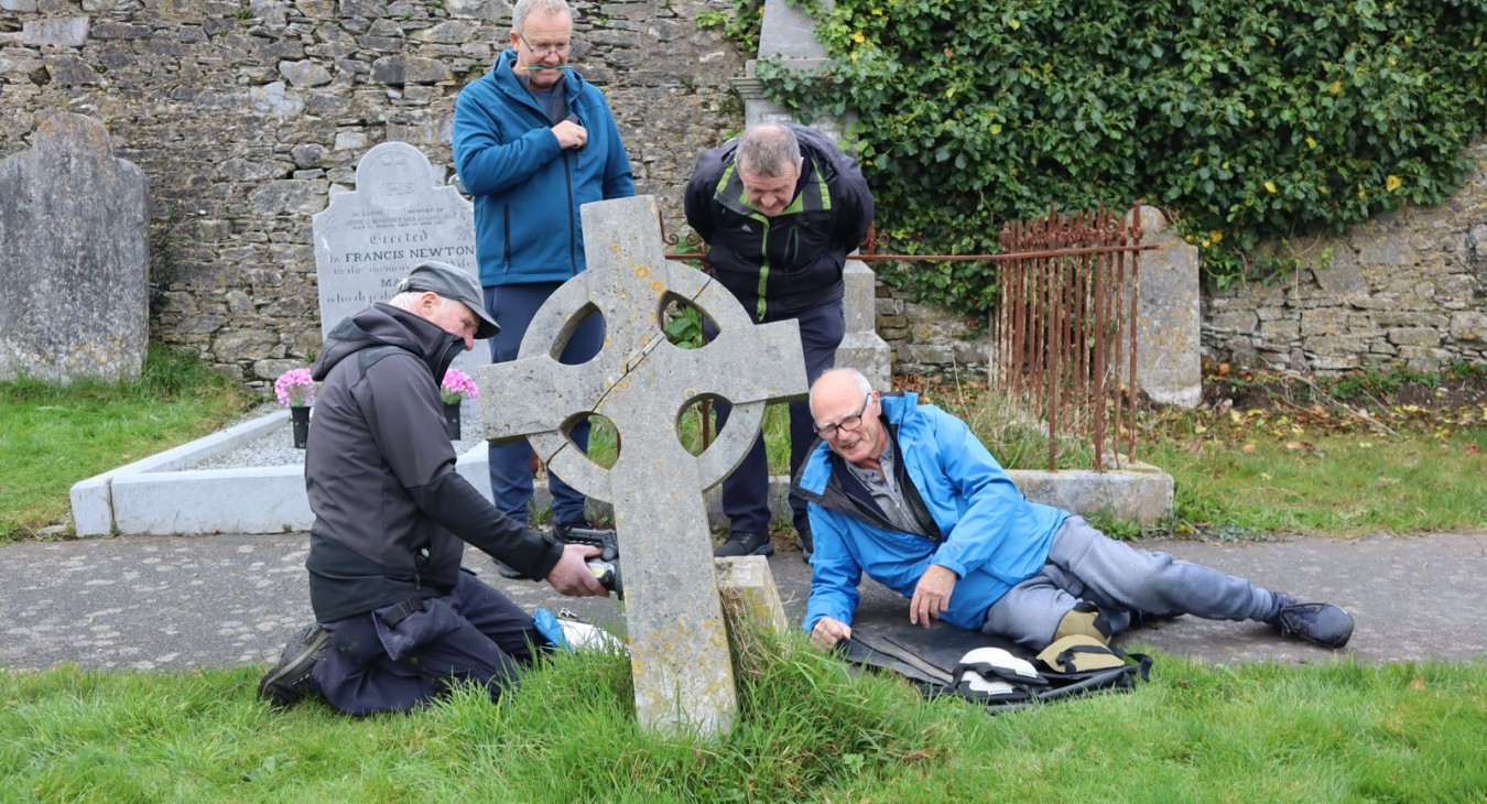Team of four men learning to record headstones; two are sitting or lying on the ground and two others stand behind them. Limestone wall in background and ringed cross in foreground.