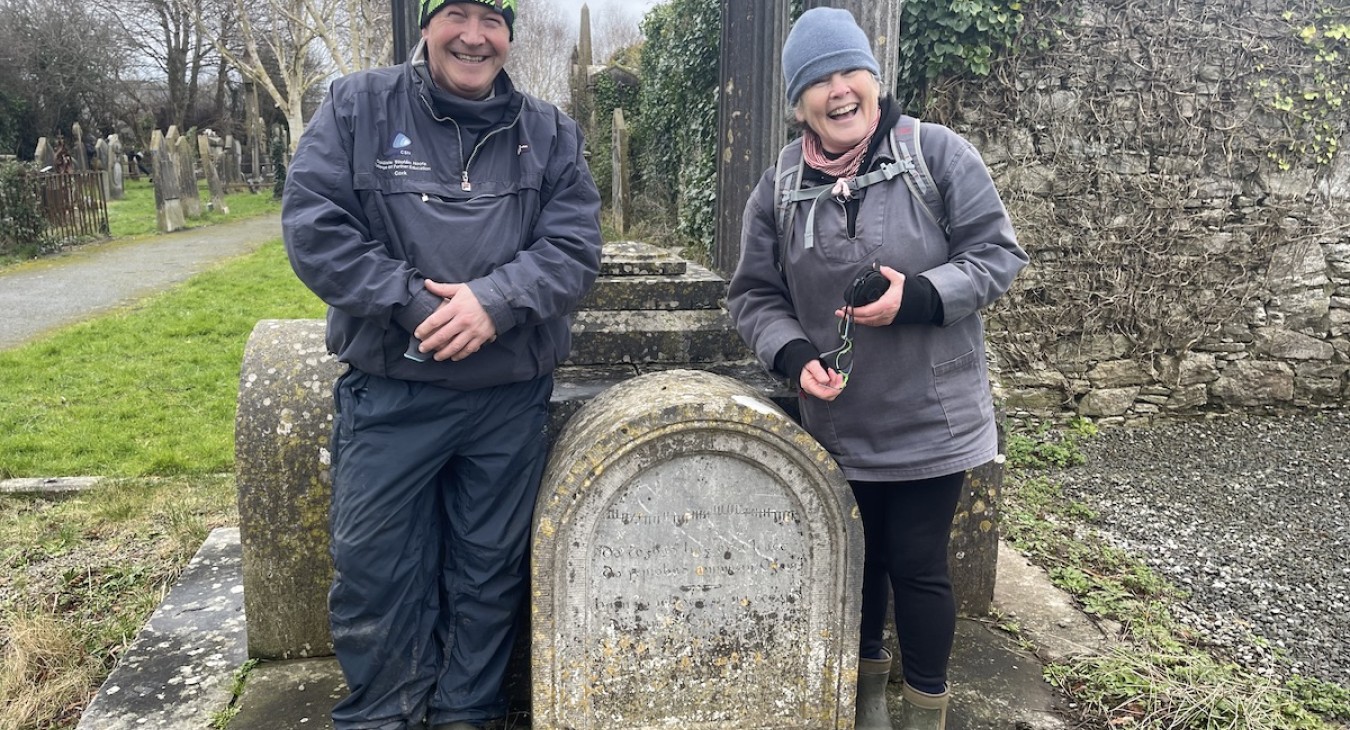 A man and woman in warm winter clothes stand in front of a large limestone grave monument in a cemetery with a large wall and adjacent pathway in background.
