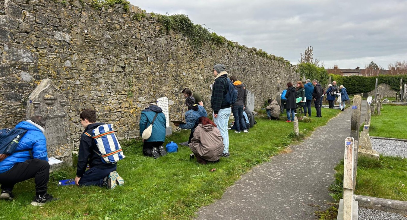 Line of students recording headstones; many on their knees on a grass surface and limestone wall behind.