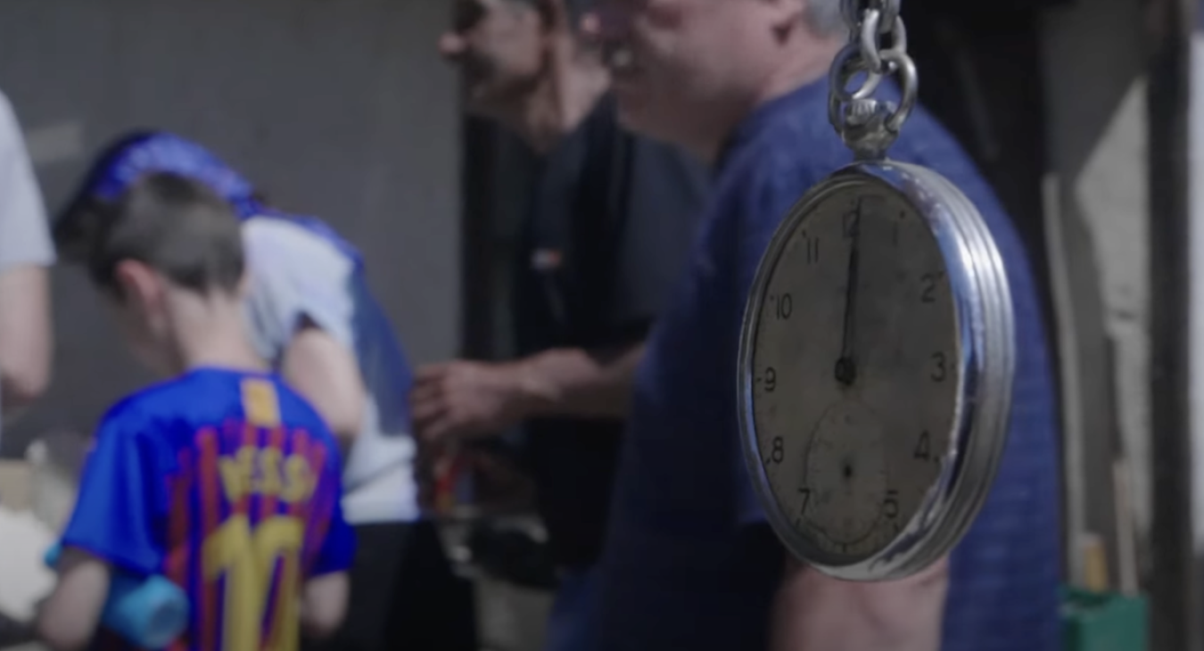 Video screencapture showing a pocket watch dangling in foreground and a boy wearing a Messi jersey in background